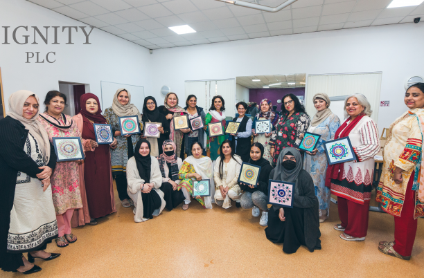 A group of women stand in a semi circle holding paper based art work. In the top left hand corner of the image, is a grey logo that reads 'Dignity PLC'